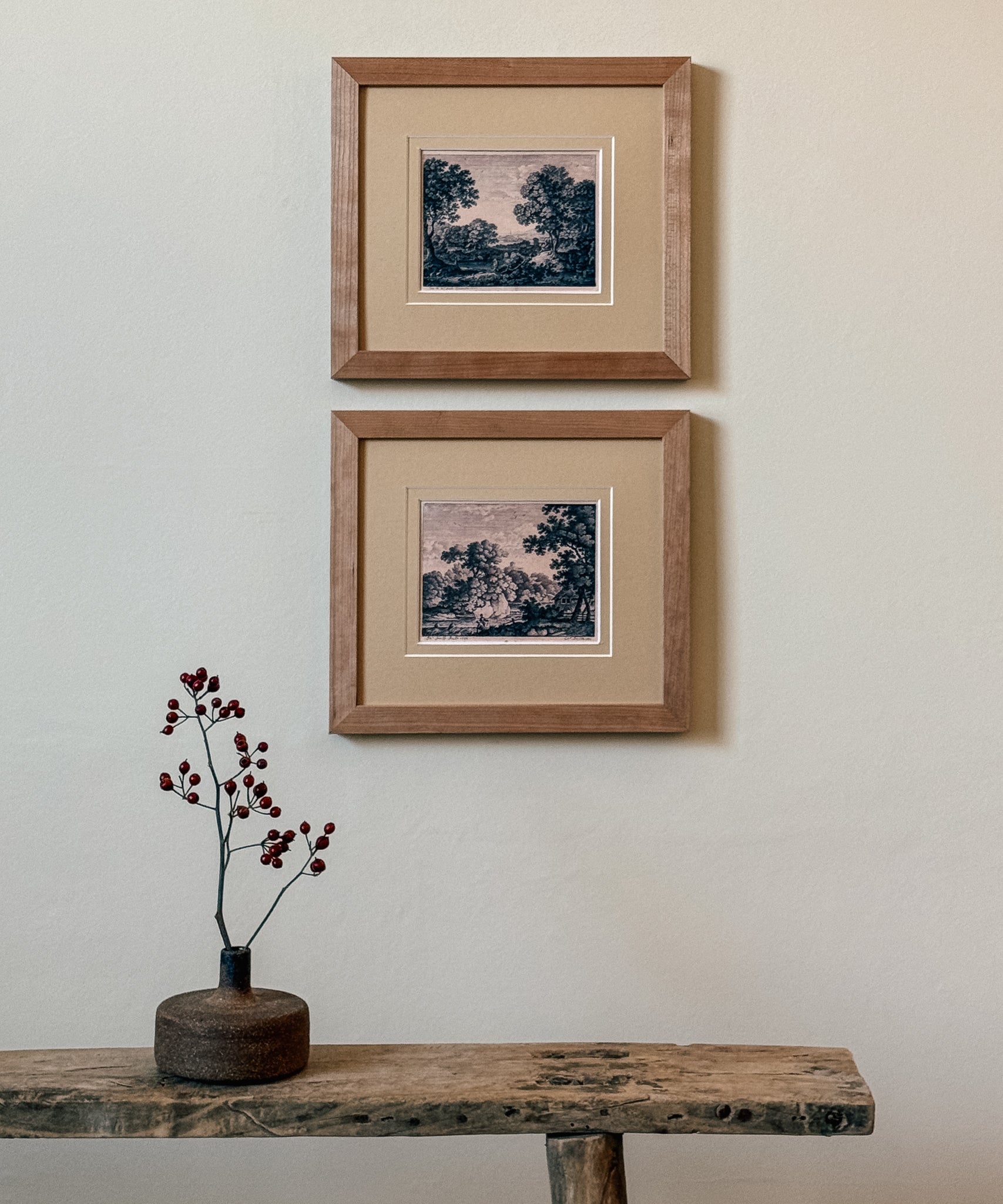 Two framed artworks on a wall above a wooden table with a vase.