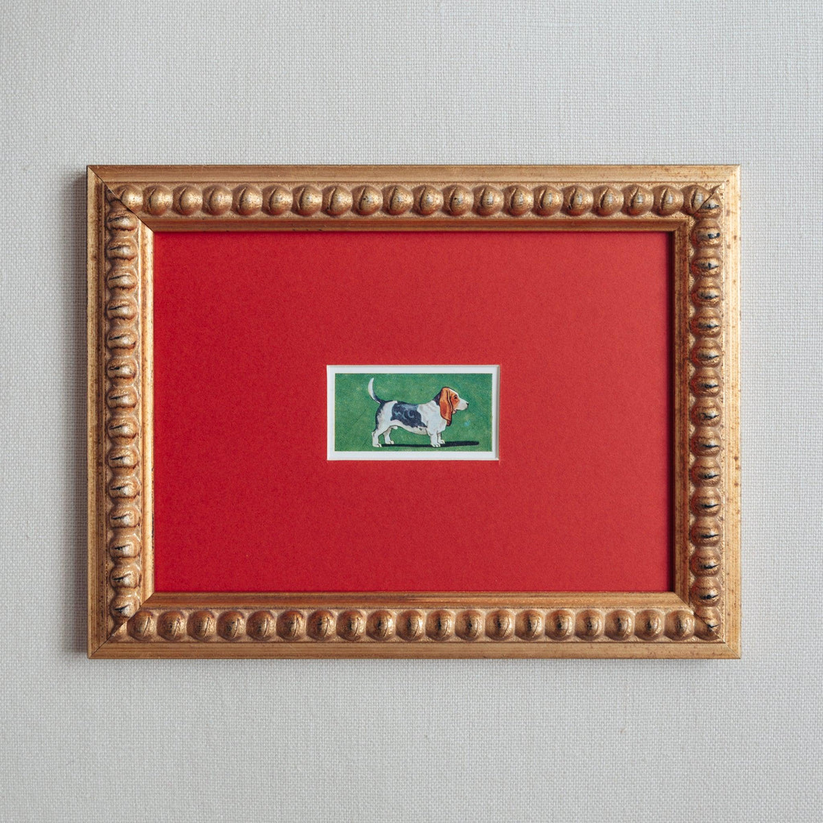 Handmade bobbin frame in antique-style gold with red mount and a simplistic lithograph of a basset hound with a green background. This has been mounted in red and is only available at Giddy Prints.